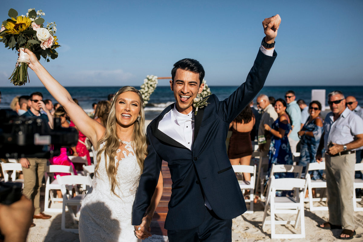 Bride and groom raise their arms and cheer walking down a beach wedding aisle with guests seated nearby