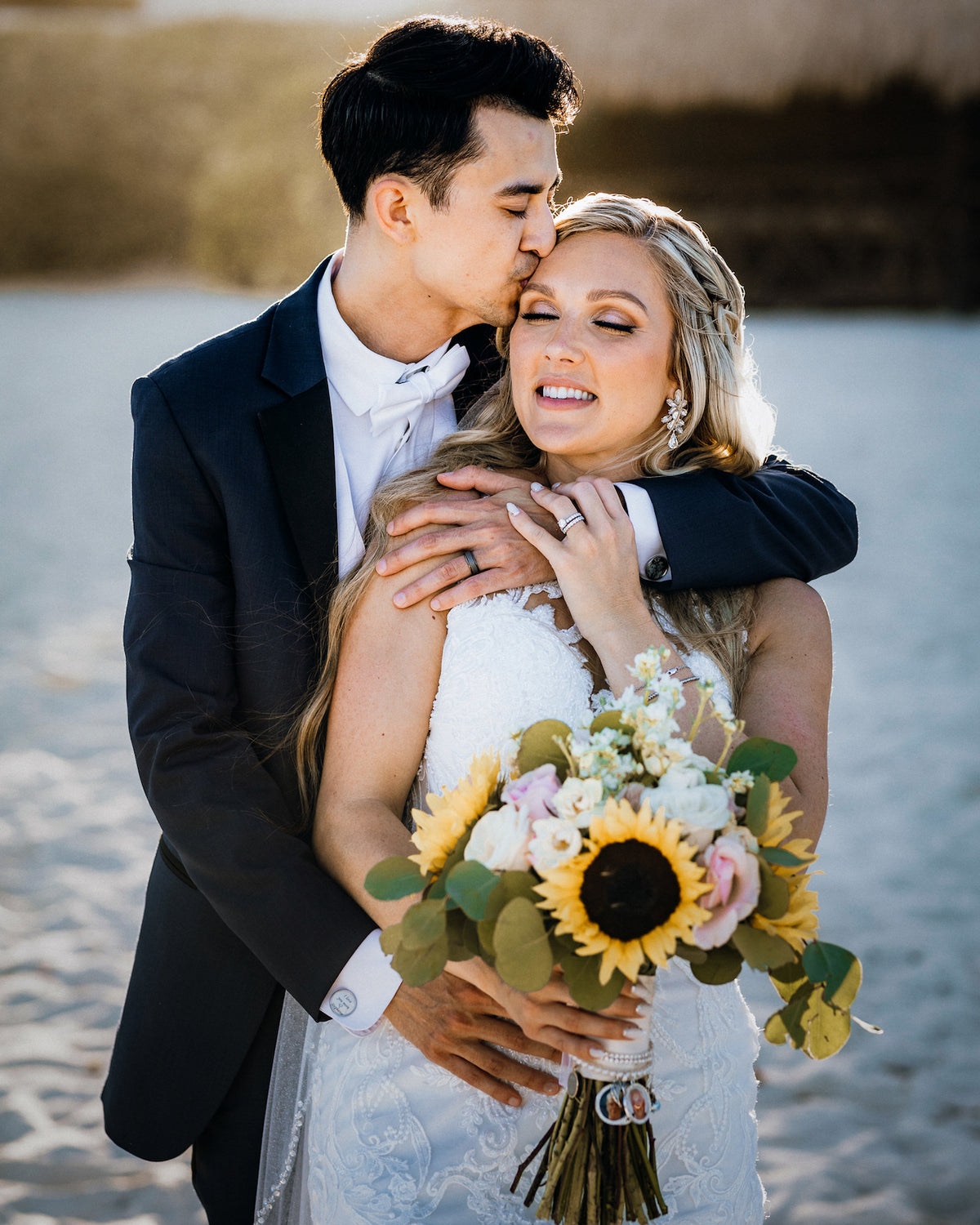 Groom in a black tuxedo kisses the forehead of a bride holding a sunflower bouquet on a beach
