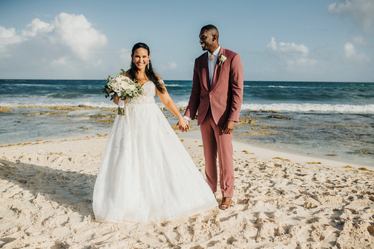 Smiling bride and groom in a rose-colored suit hold hands on a white sand beach wedding
