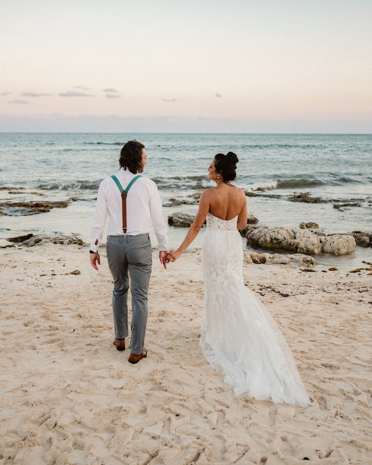 Bride and groom walk hand in hand across the sand toward the ocean at sunset