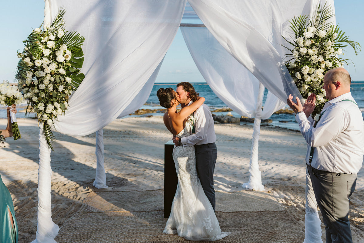 Bride and groom share their first kiss under a white draped beach canopy as a guest applauds