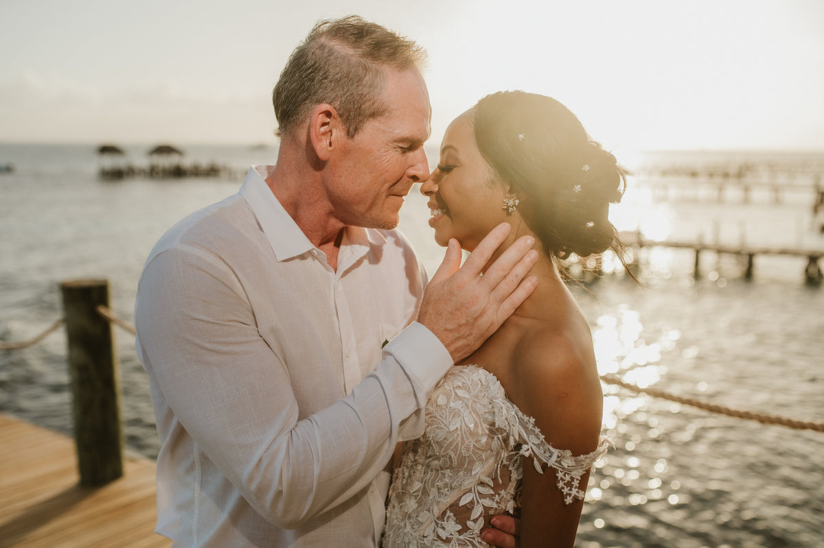 Groom cradles his bride's face as they touch noses on a sunlit pier by the sea