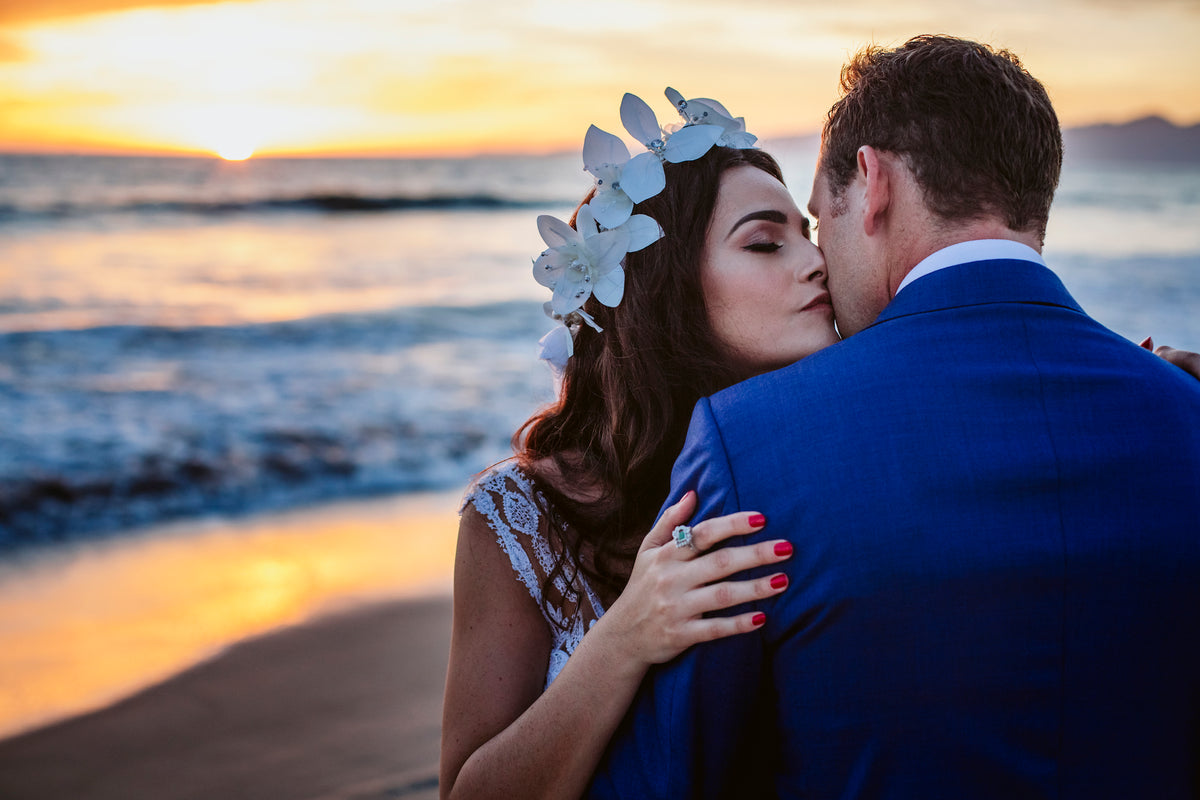 Bride with orchids in her hair kisses the groom in a blue suit on the beach at sunset