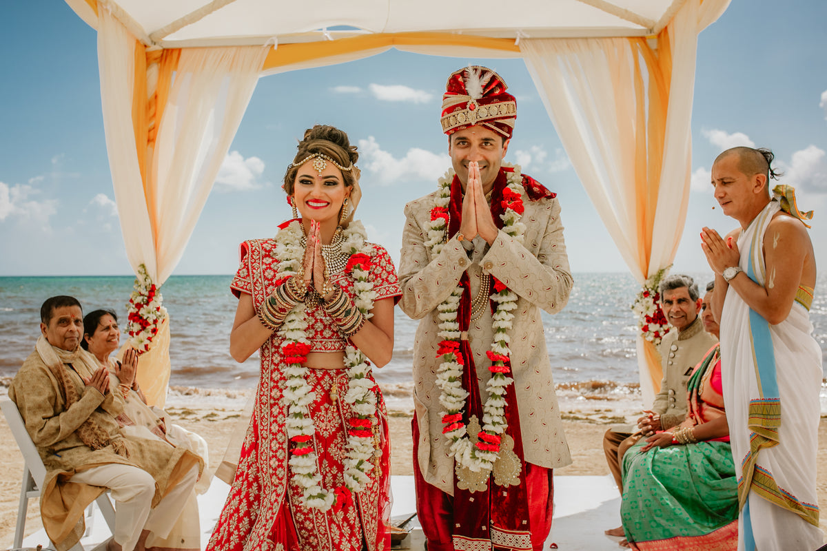 Bride and groom in traditional Indian wedding attire with hands folded under a beach canopy at Moon Palace Cancún