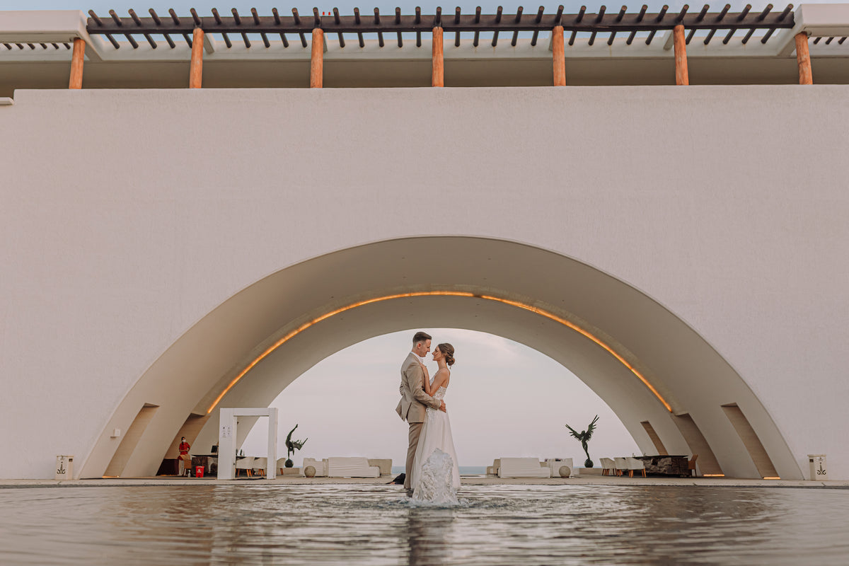 A bride and groom embrace beneath a large stone archway at Marquis Los Cabos, standing in a pool