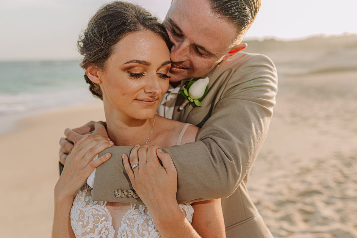 Bride and groom embrace on a beach, ocean visible behind them