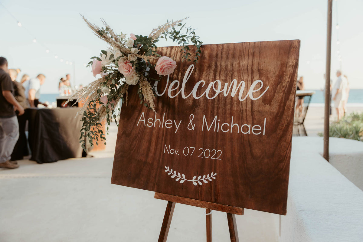 Wooden welcome sign for Ashley and Michael decorated with flowers, displayed at a beachside wedding at Marquis Los Cabos