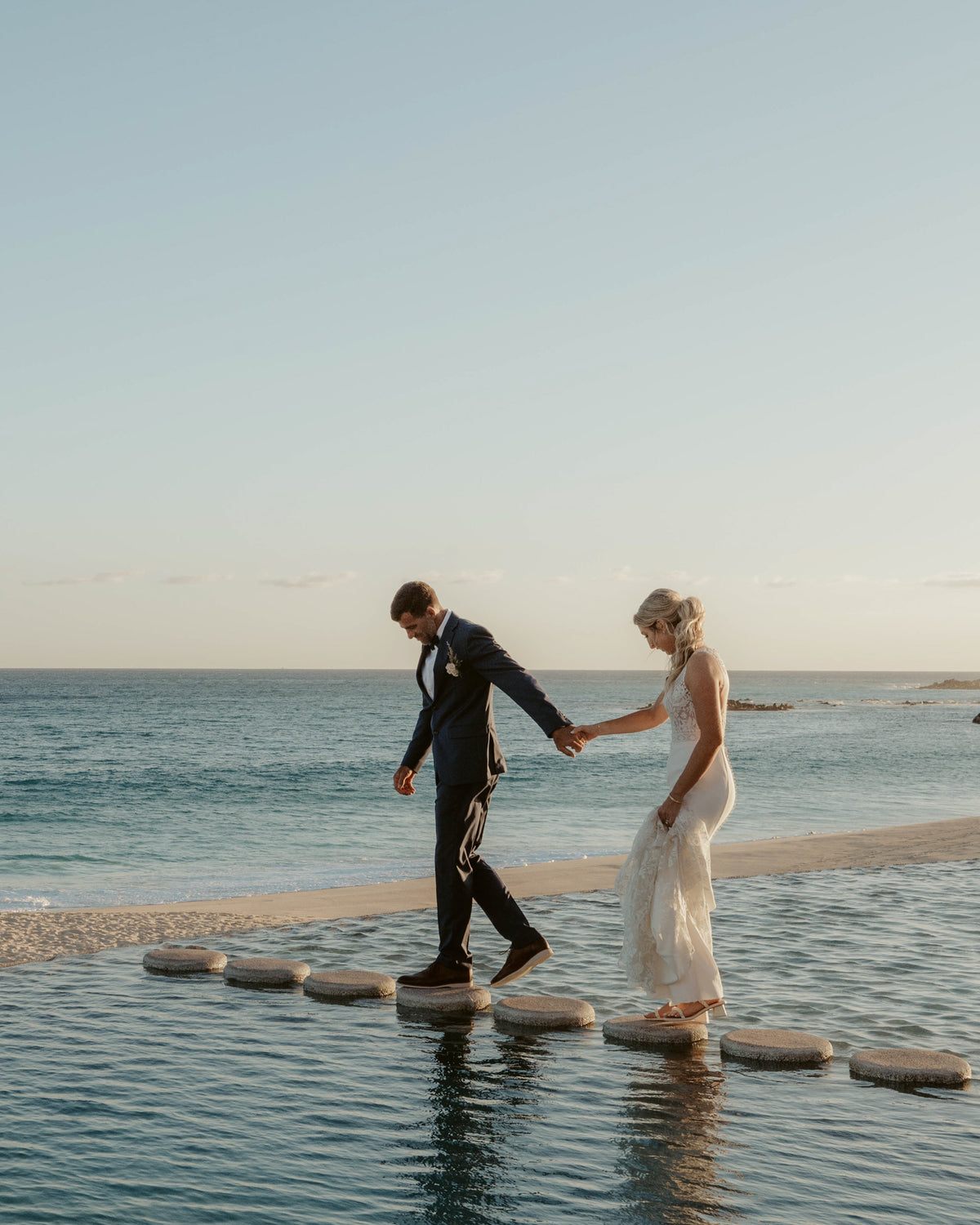 Bride and groom cross stone stepping pads over water at Marquis Los Cabos, ocean behind them