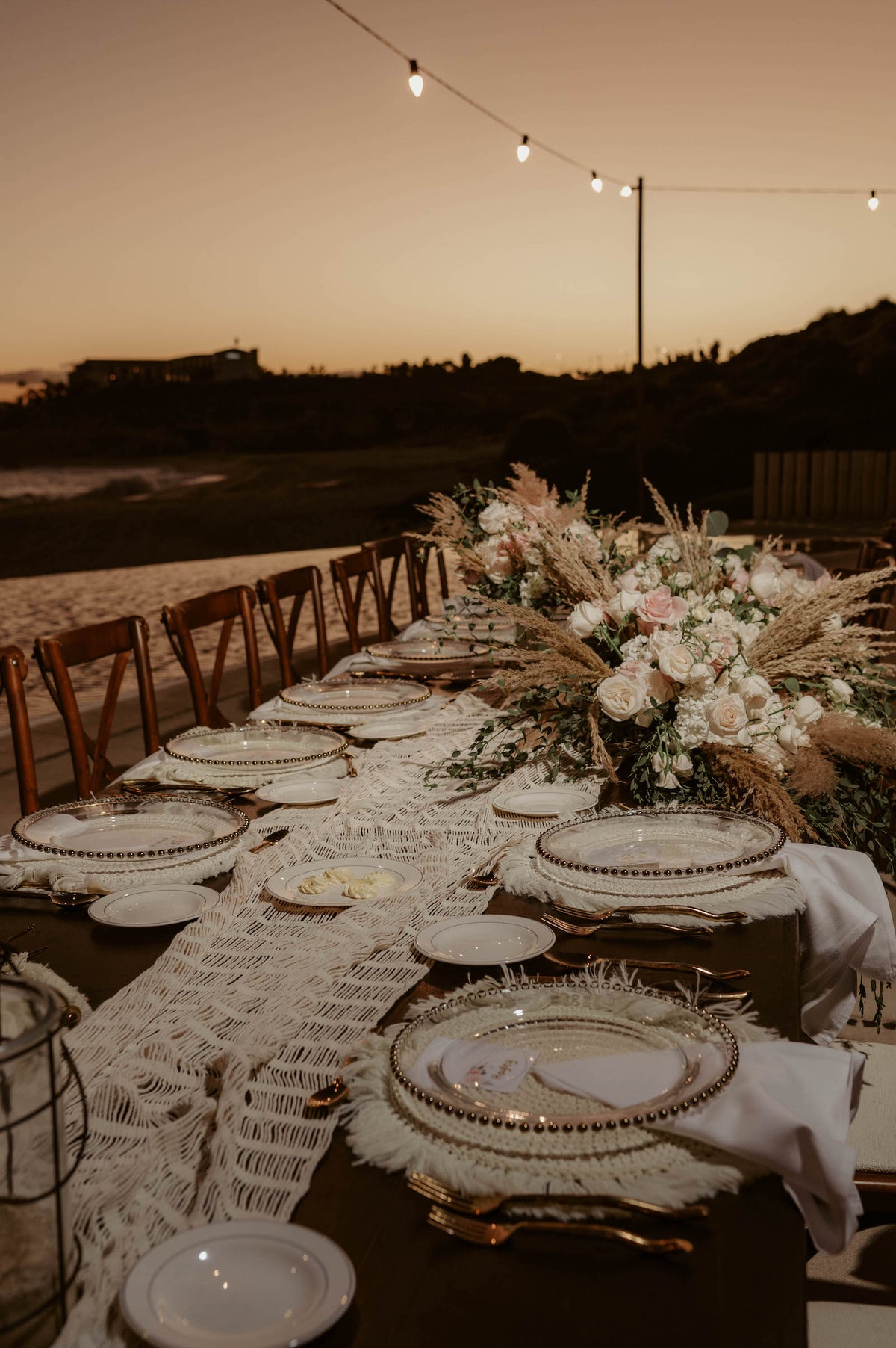 Outdoor wedding reception table with macrame runner, floral centerpiece, and string lights at Marquis Los Cabos during sunset