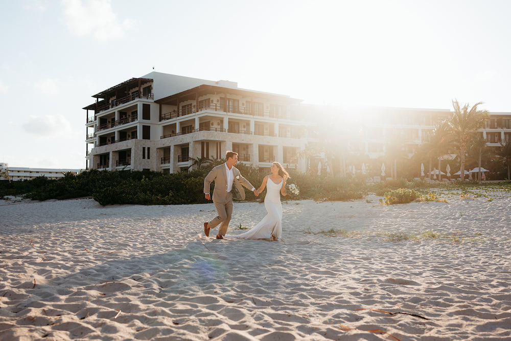 A bride and groom hold hands on a beach at Secrets Playa Blanca in Costa Rica, resort buildings behind them