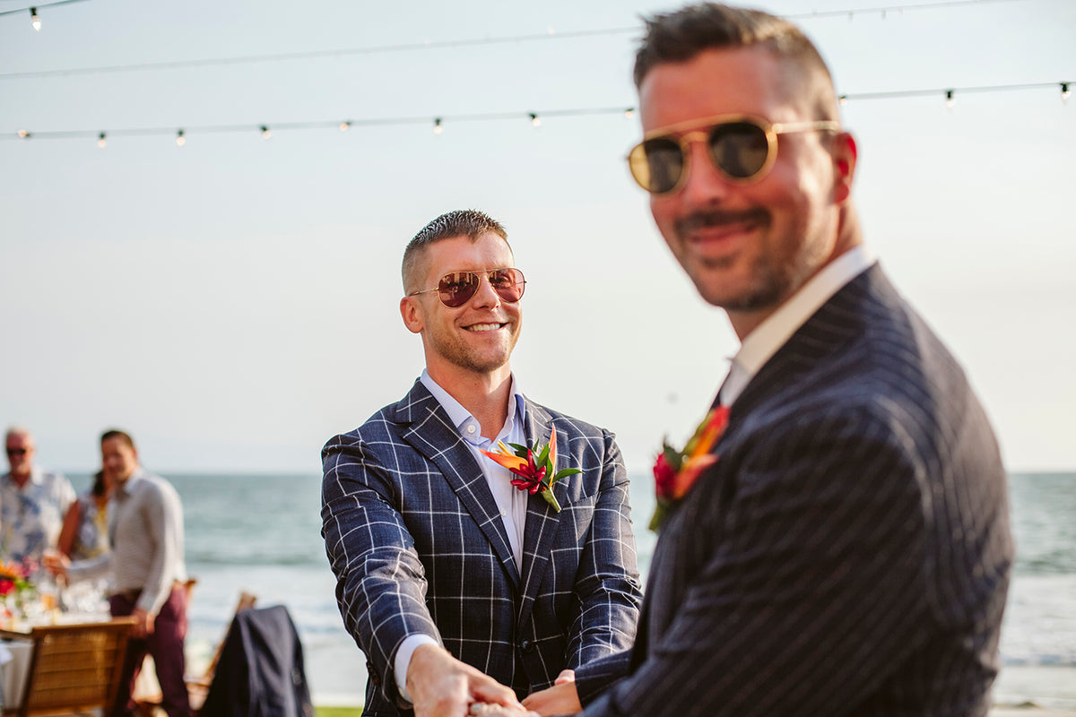 Two grooms with tropical boutonnieres hold hands and smile during their beachfront wedding celebration