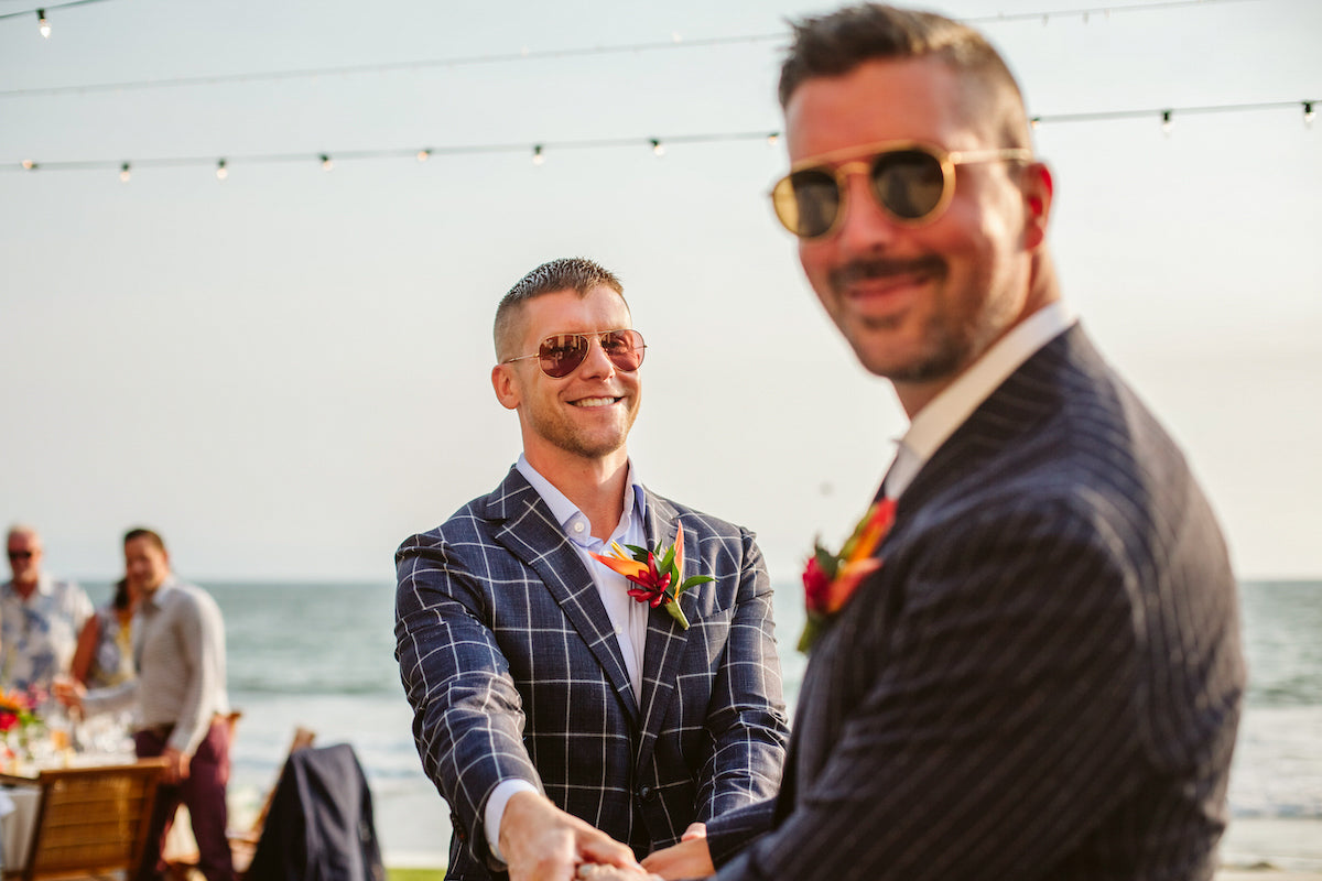 Smiling groom in aviator sunglasses holds his partner's hands at a seaside same-sex wedding