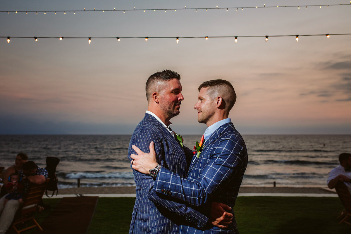 Two grooms in blue suits share a dance by the ocean under string lights at dusk