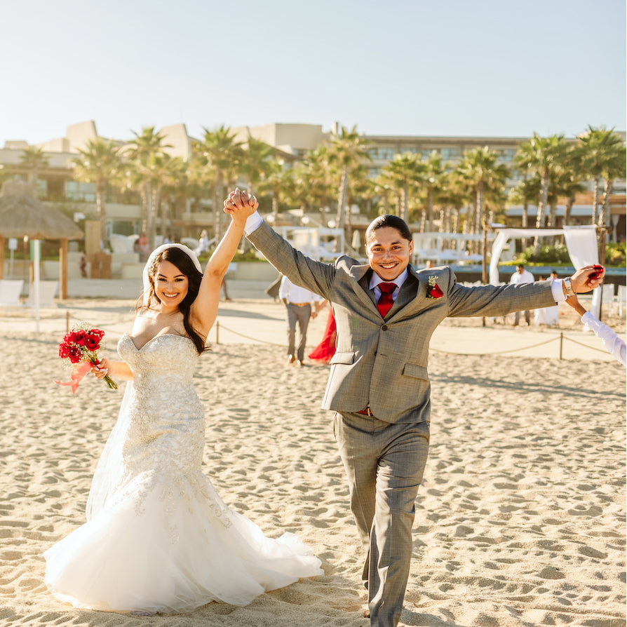 A bride and groom raise joined hands while walking across a beach near palm trees and a resort building
