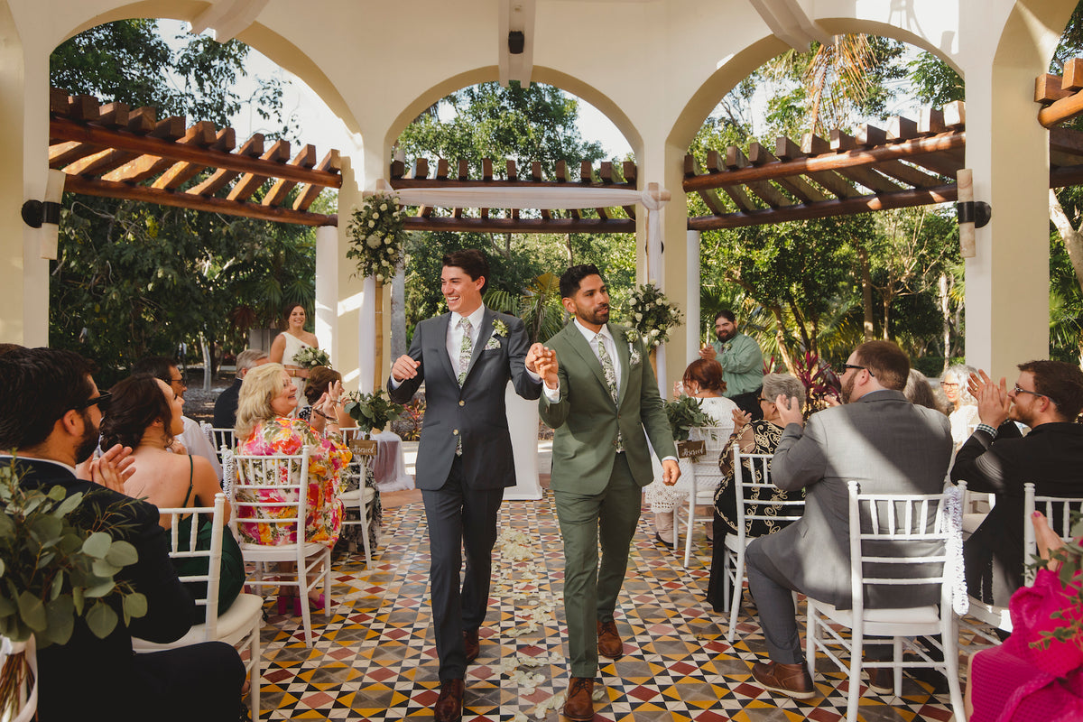 Two grooms walk hand in hand down the aisle as guests applaud at Valentin Imperial Riviera Maya