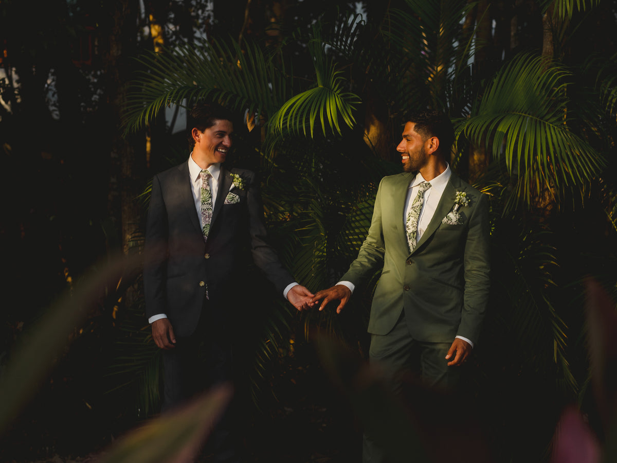 Two grooms in suits reach for each other's hands among palm fronds at Valentin Imperial Riviera Maya