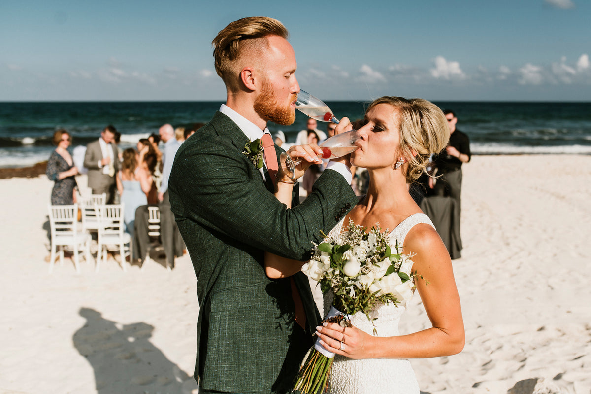 Bride and groom link arms drinking champagne on the beach at Valentin Imperial Riviera Maya