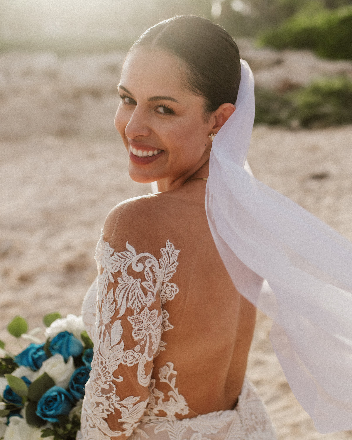 A bride with a lace-sleeved gown and veil smiles over her shoulder on the beach at Valentin Imperial Riviera Maya