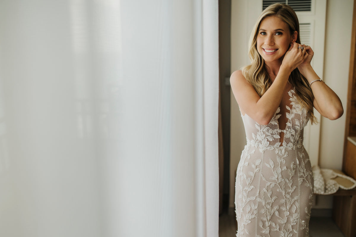 Bride in a lace gown fastening an earring by a bright window while getting ready at JW Marriott Cancun