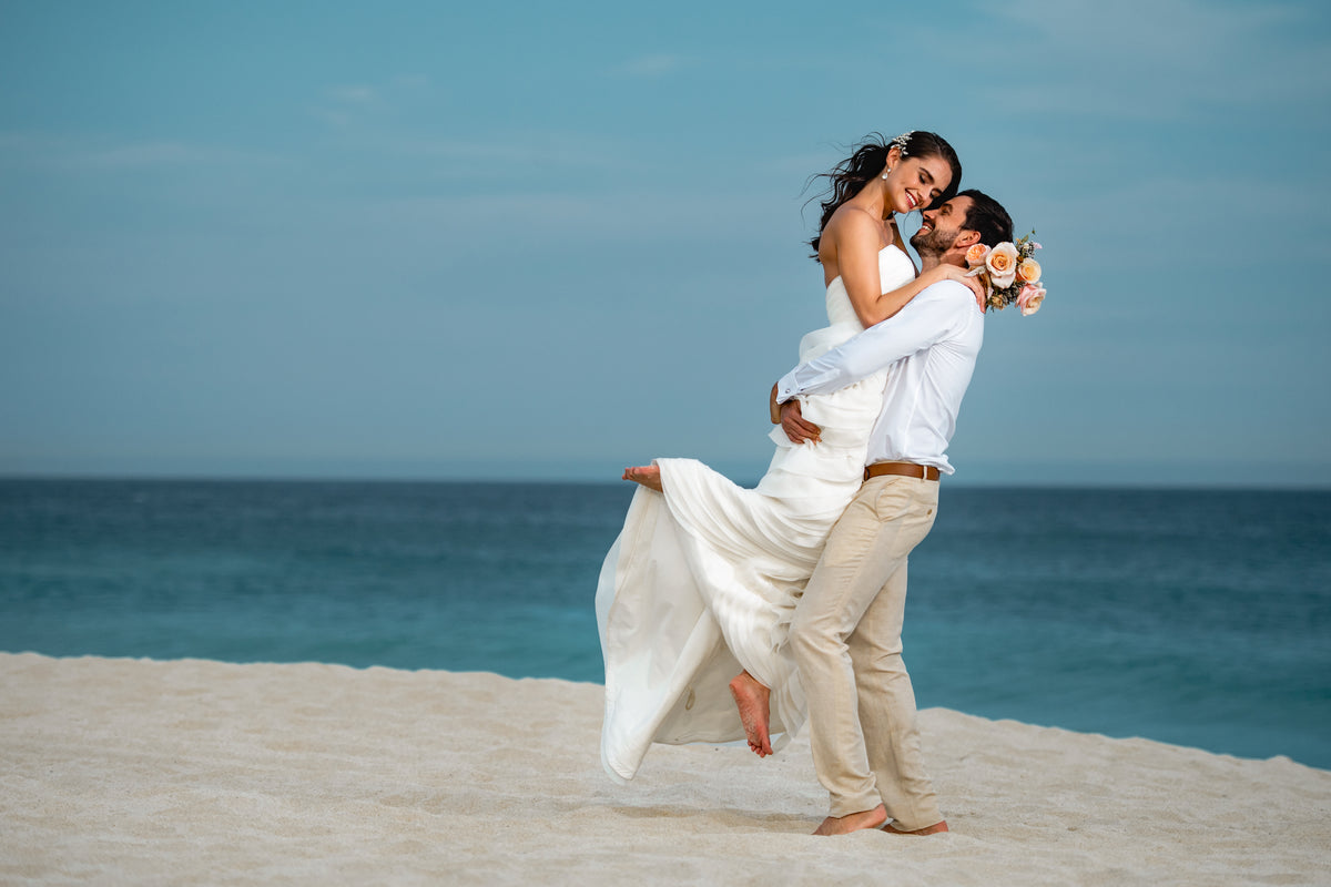 Groom lifting the bride in his arms on a sandy beach with the ocean behind