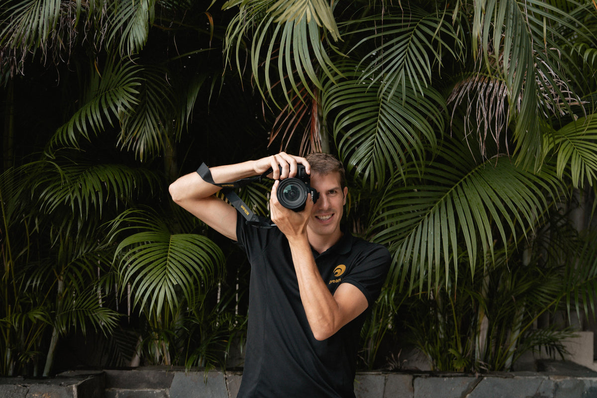 Smiling DreamArt photographer in a black polo aims his camera in front of tropical palm leaves