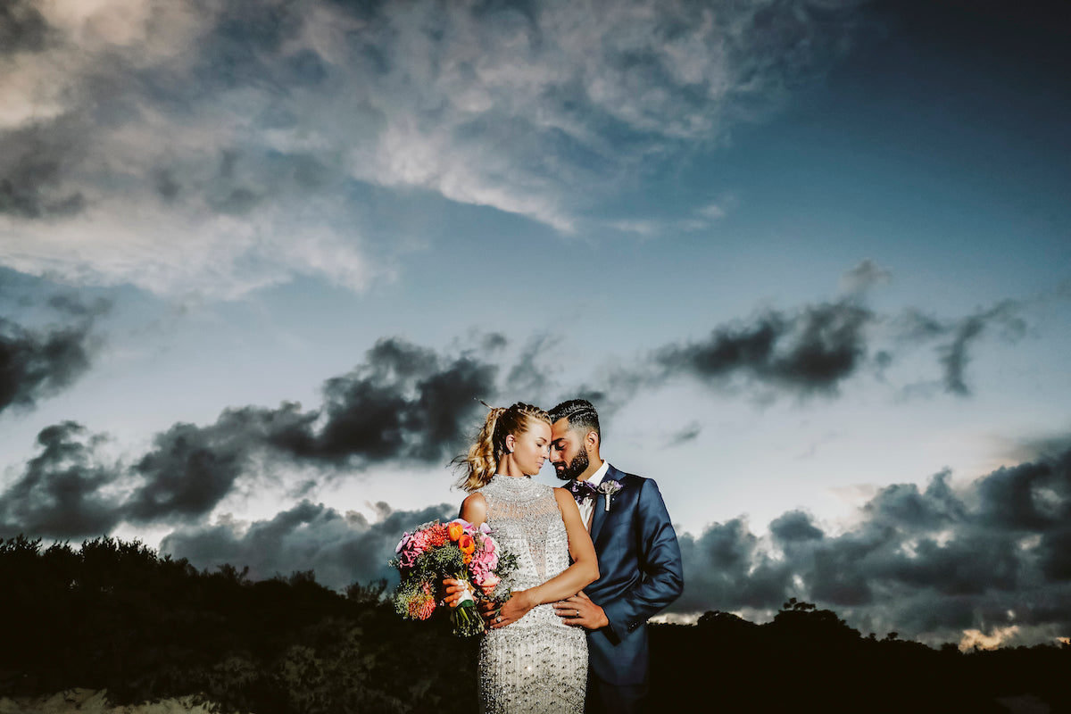 Bride with a colorful bouquet and groom touch heads under a dramatic twilight sky at Banyan Tree Mayakoba