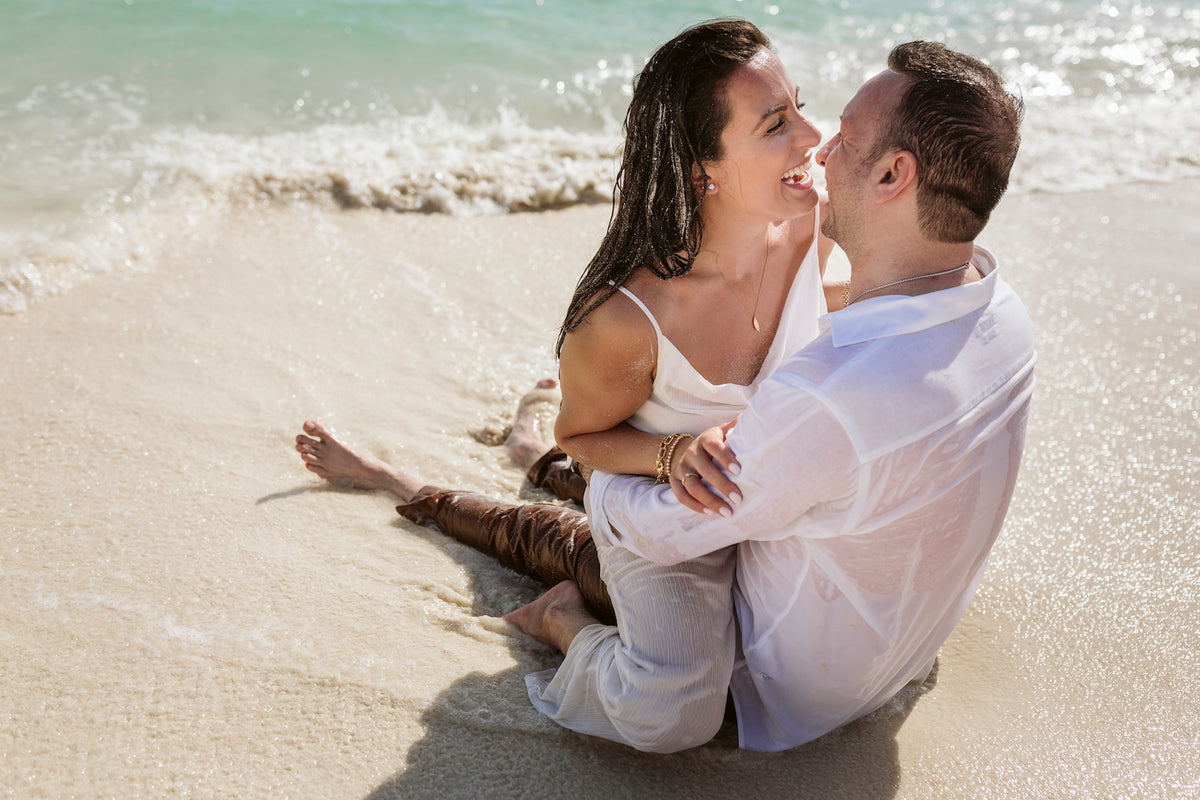 Laughing couple in white sit together in the gentle surf during a beach photoshoot in Cancun