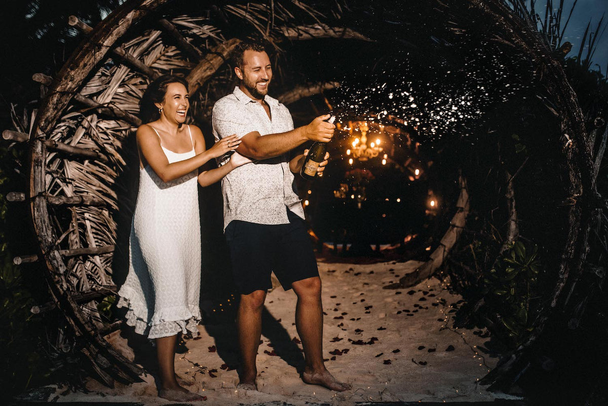 Couple celebrate their engagement by spraying champagne under a woven branch arch at night at Casa Malca