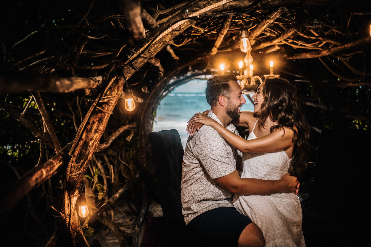 Woman sits on her partner's lap laughing under a candle chandelier in a branch tunnel facing the sea