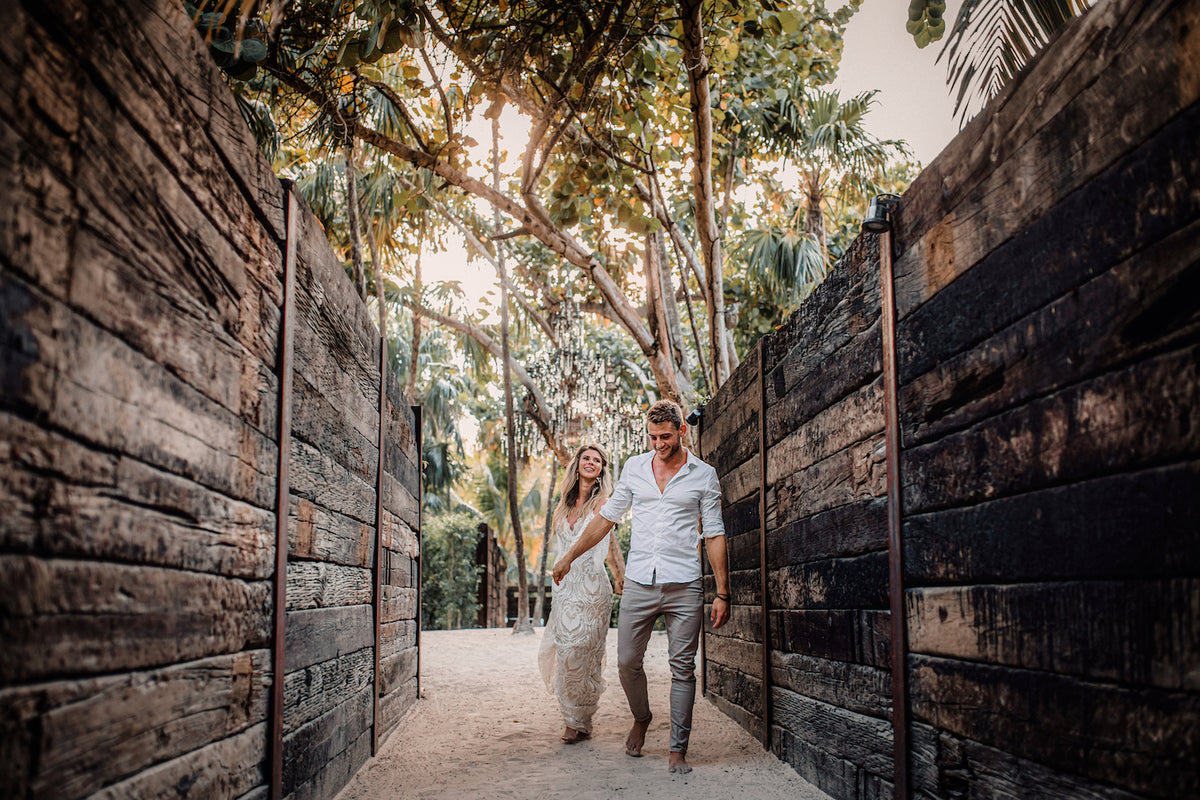 Bride and groom stroll through a dark timber corridor toward tropical trees at Casa Malca