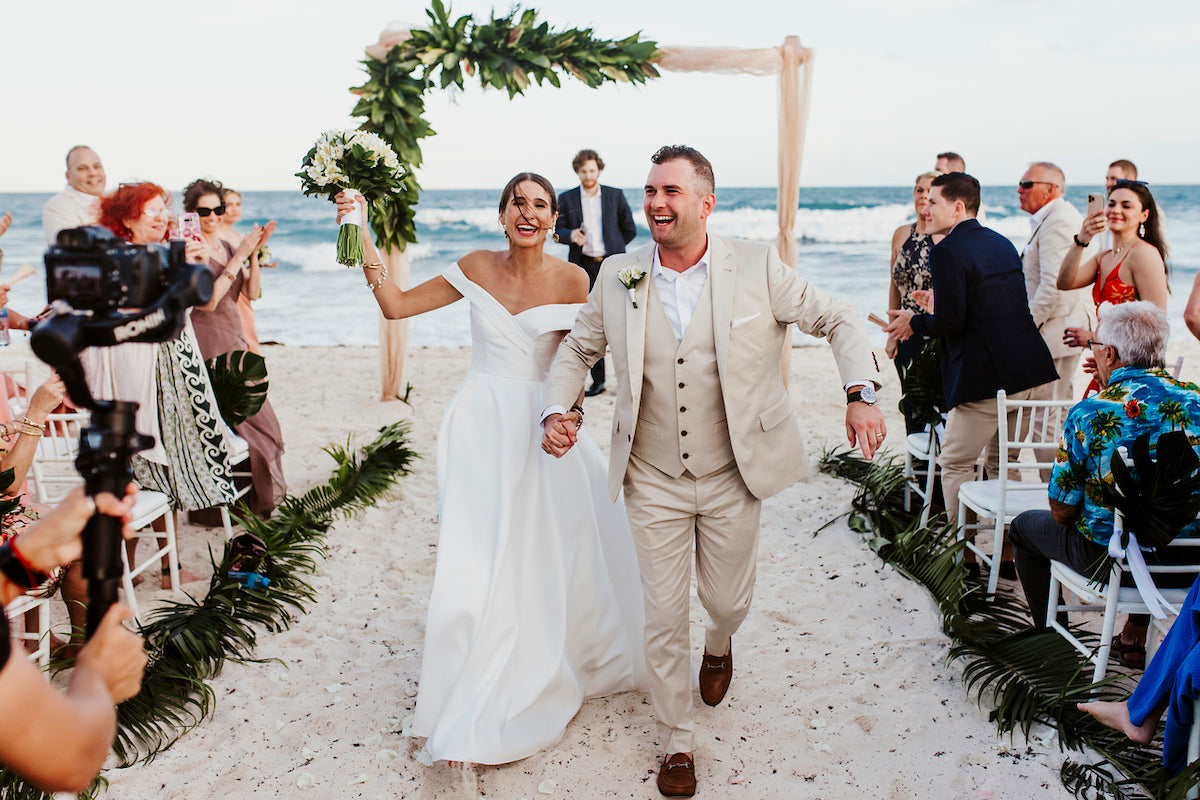 Joyful bride and groom walk back down a palm-lined beach aisle as guests cheer after the ceremony