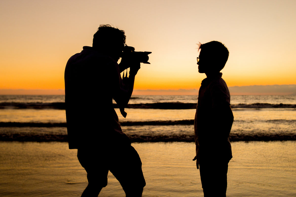 Silhouette of a photographer shooting a boy in glasses on the beach at sunset