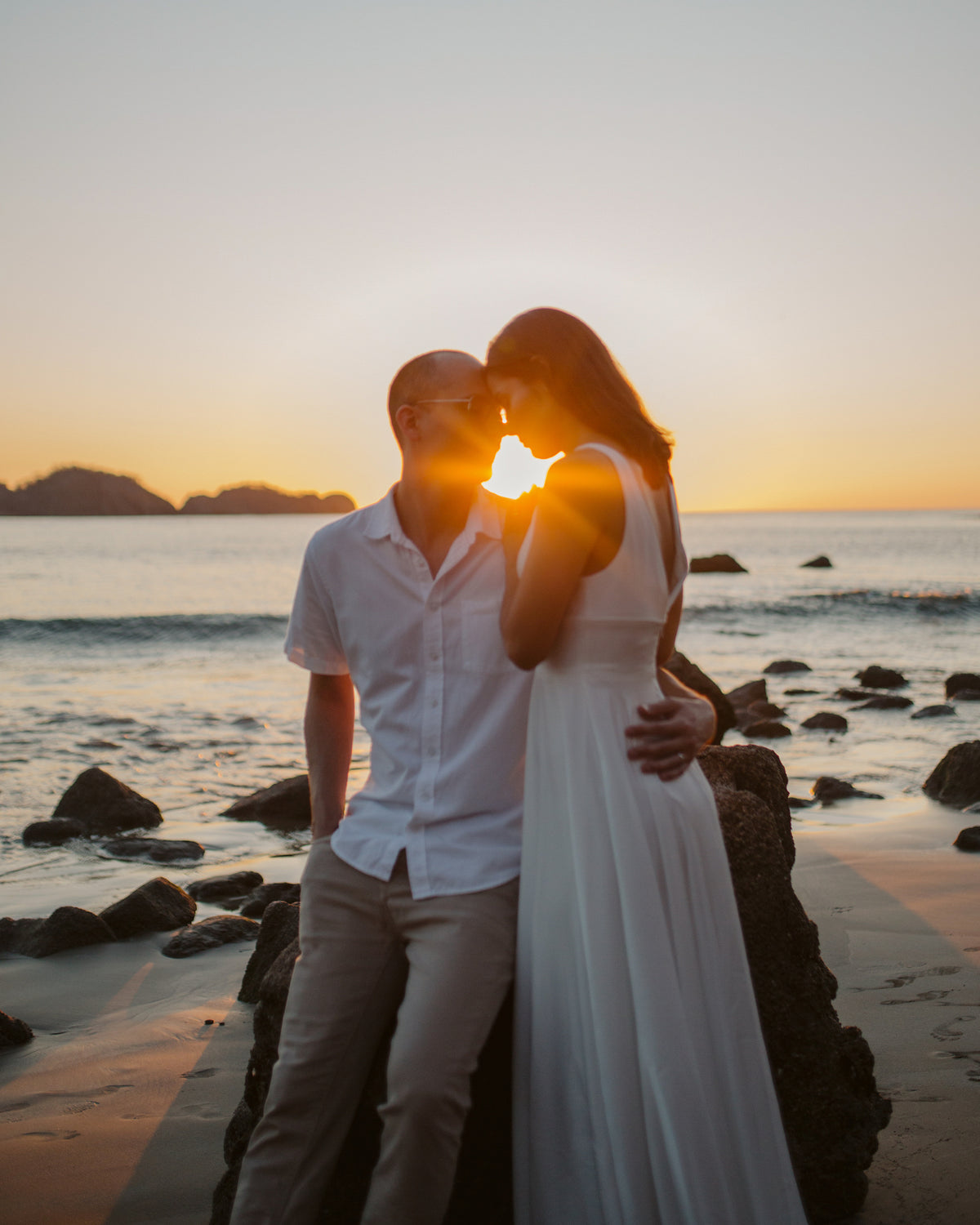 Couple touching foreheads on beach rocks as the sun sets behind them in Costa Rica