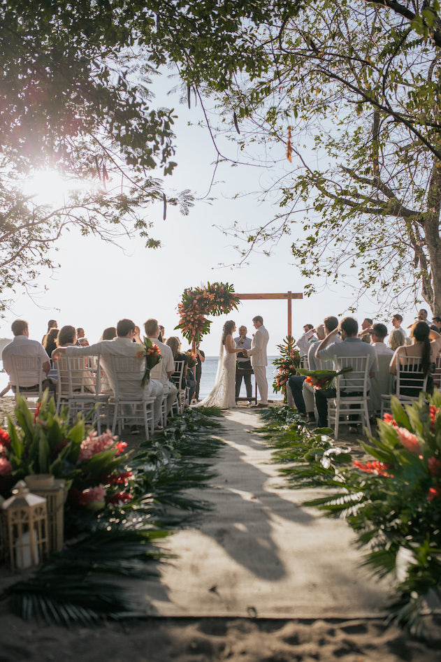 Beachfront wedding ceremony beneath trees with a tropical flower arch and palm-lined aisle in Costa Rica