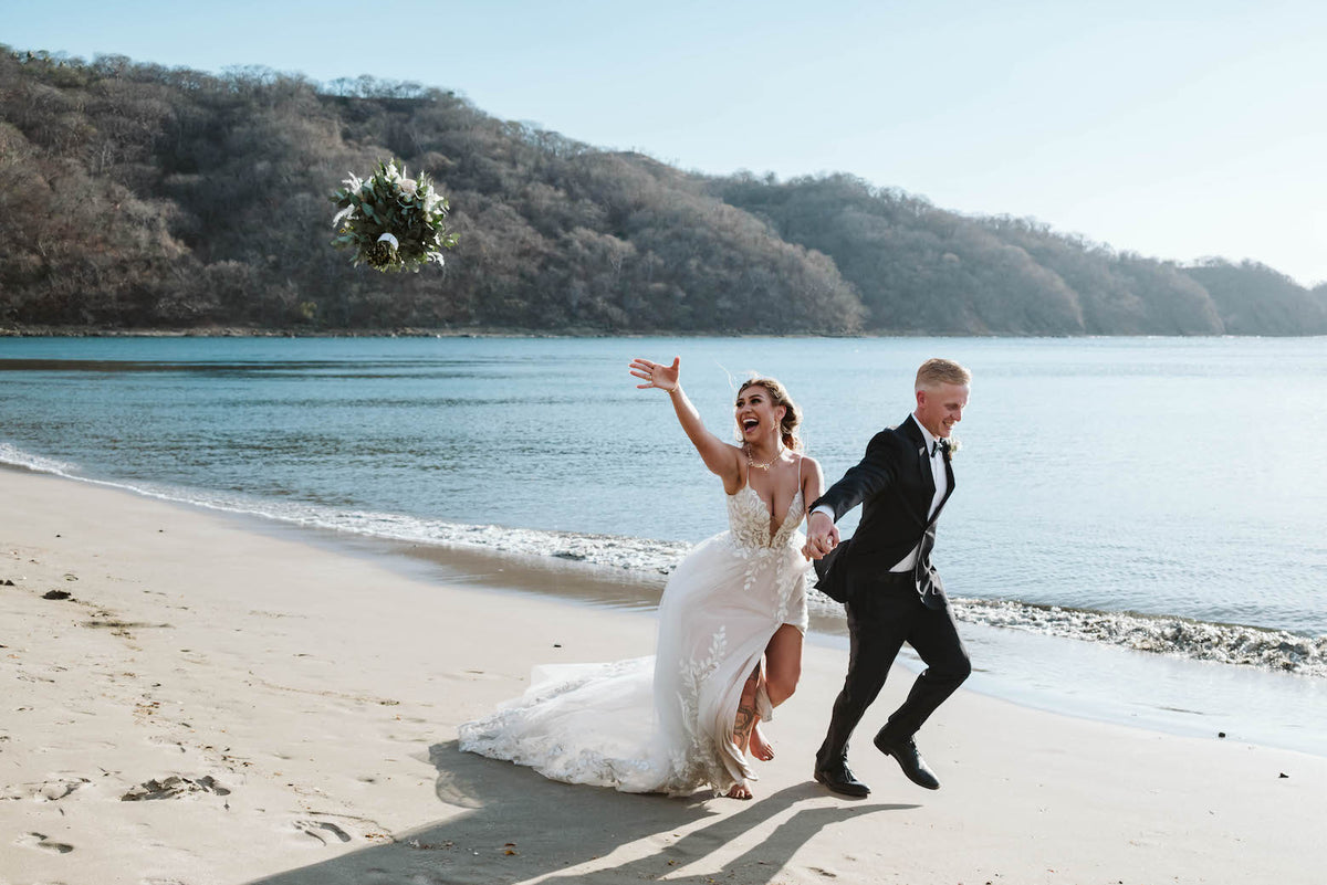 Bride tosses her bouquet into the air as she runs along the beach with the groom in Costa Rica