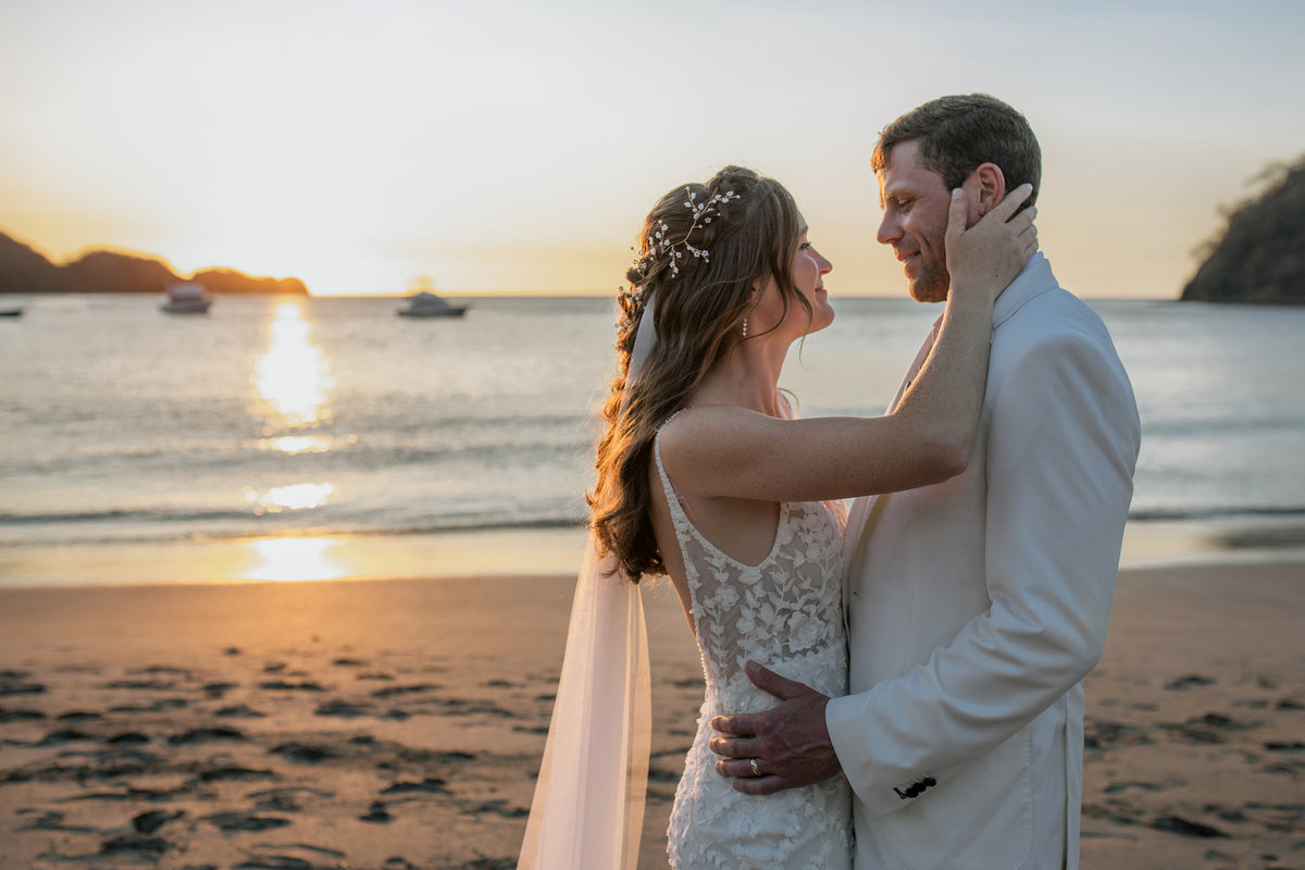 Bride touches the groom's face on the beach at sunset with boats on the bay in Costa Rica