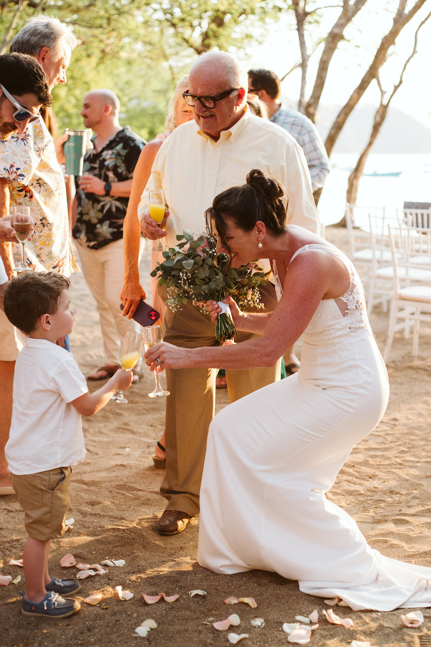 Bride kneels to clink glasses with a little boy during a beach wedding celebration in Costa Rica