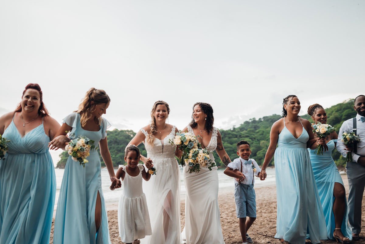Two brides walk arm in arm on the beach with bridesmaids in pale blue and two children in Costa Rica