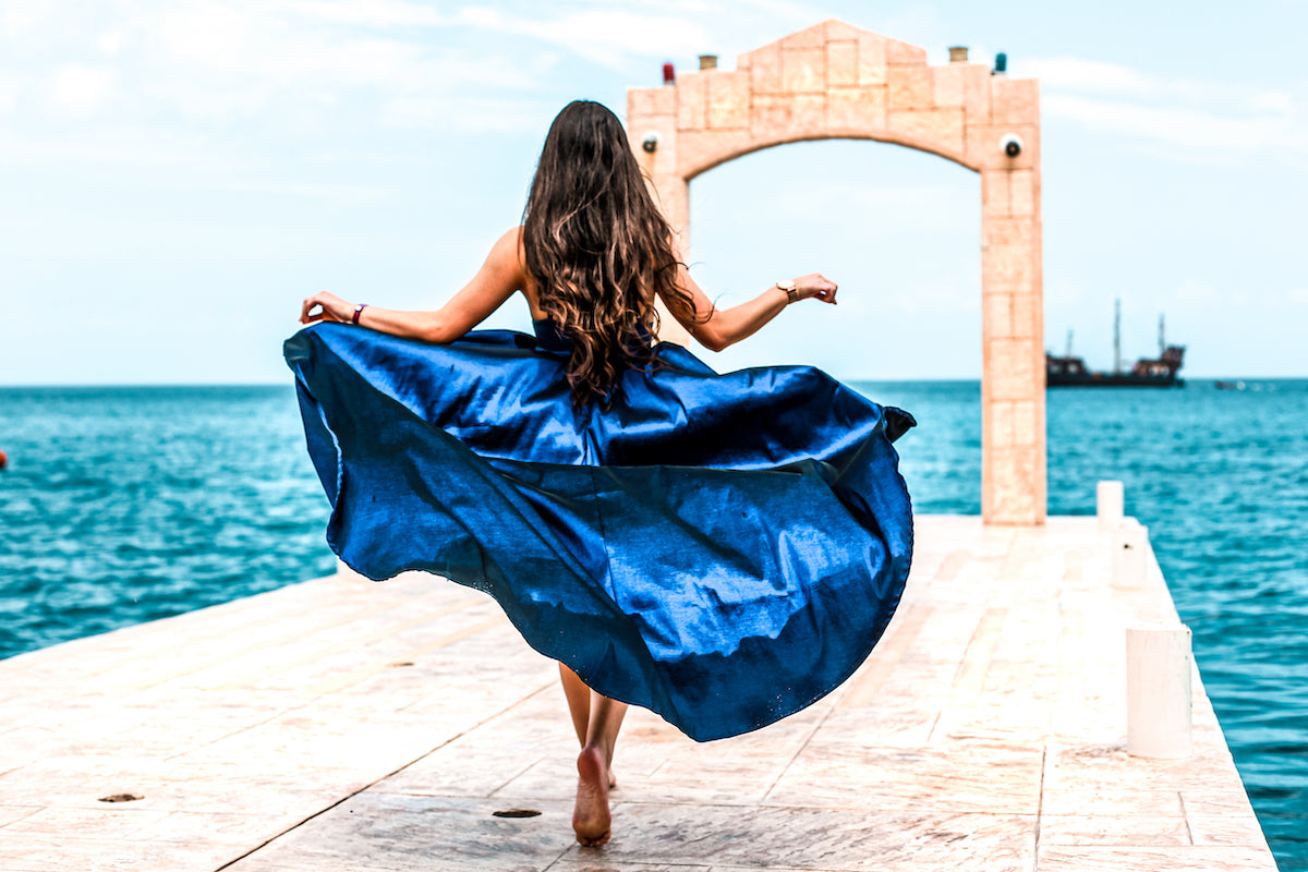 Woman in a flowing blue gown walks a pier toward a stone arch over the sea at Cozumel Palace