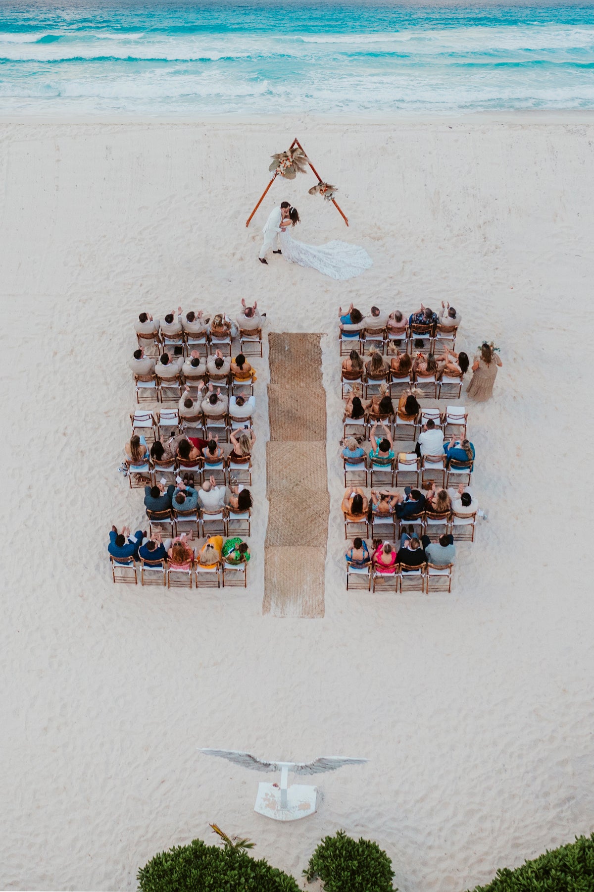 Drone view of a beach wedding ceremony with guests watching the couple kiss under a boho arch