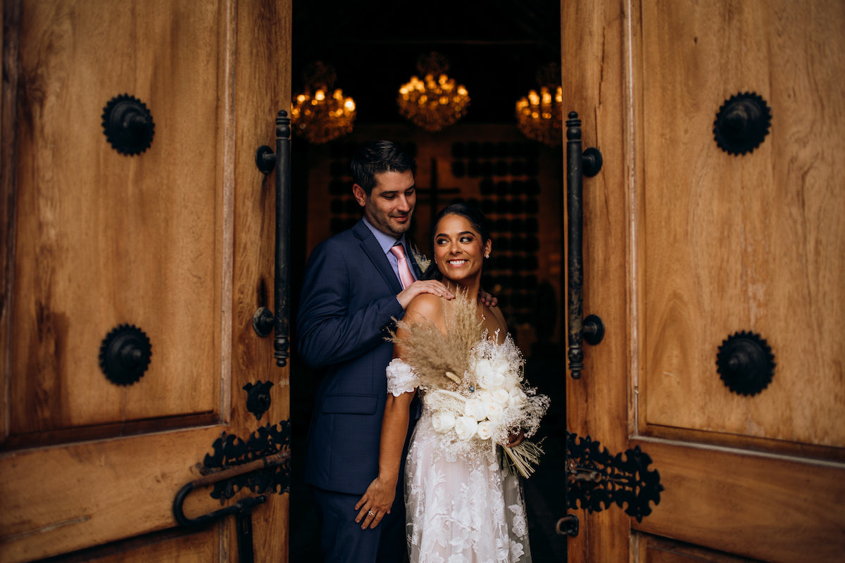 Bride and groom pose between carved wooden doors with chandeliers glowing behind at Fairmont Mayakoba