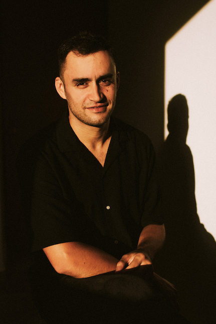 Portrait of a bootcamp instructor in a black shirt seated in warm window light, his shadow on the wall