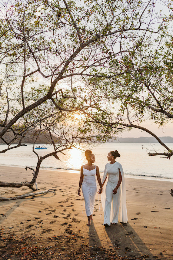 Two brides walk hand in hand along the beach beneath a tree at sunset in Guanacaste