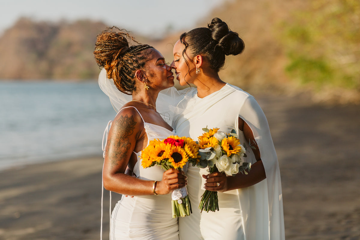 Two brides in white touch noses while holding sunflower bouquets at their Guanacaste beach wedding