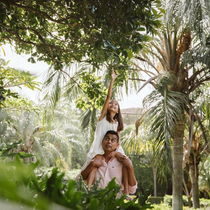 Girl reaches up to the trees from her father's shoulders in a tropical garden in Guanacaste