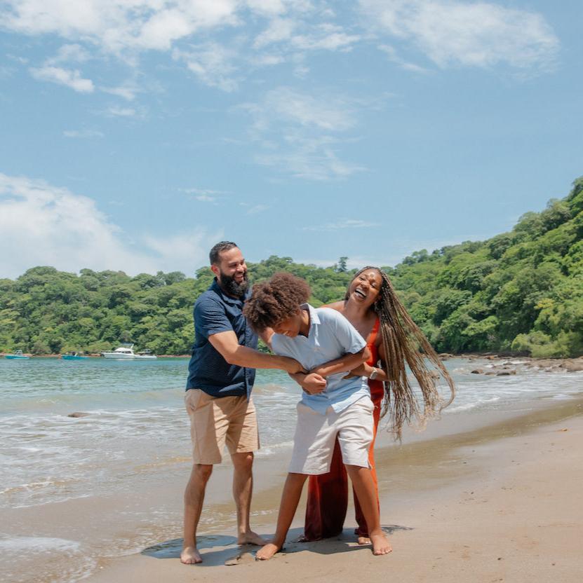 Father and son play-wrestle as mom laughs on a jungle-backed beach in Guanacaste, Costa Rica