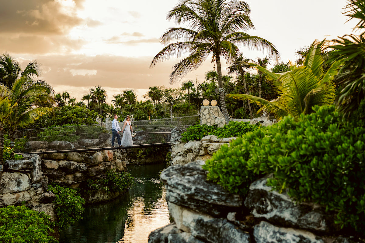 Bride and groom cross a stone bridge over a jungle river at sunset at Hotel Xcaret
