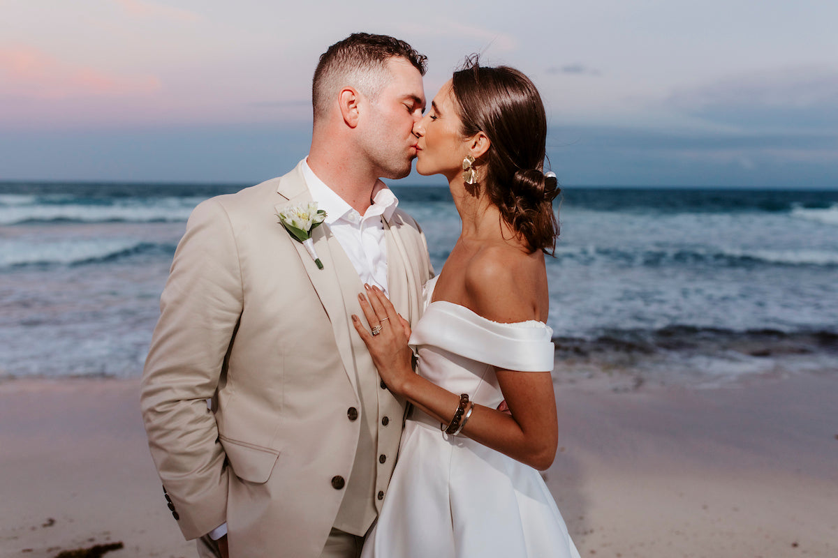 Bride in an off-shoulder gown kisses the groom in a beige suit on the beach at dusk