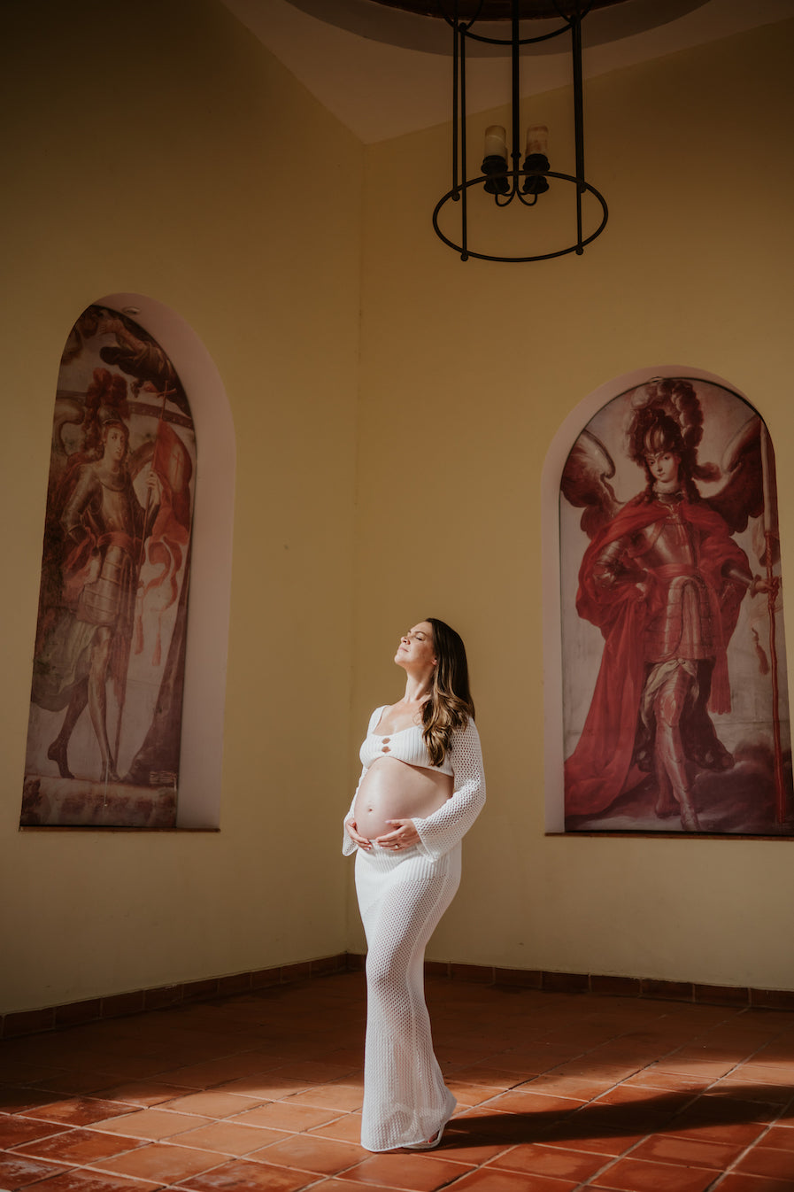 Pregnant woman in a white knit dress bathed in sunlight in a hall with painted arches during a maternity photoshoot