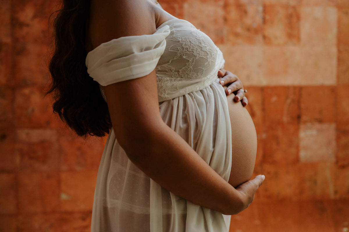 Expecting mother in a white lace off-shoulder dress cradles her bump against a terracotta wall during a maternity photoshoot